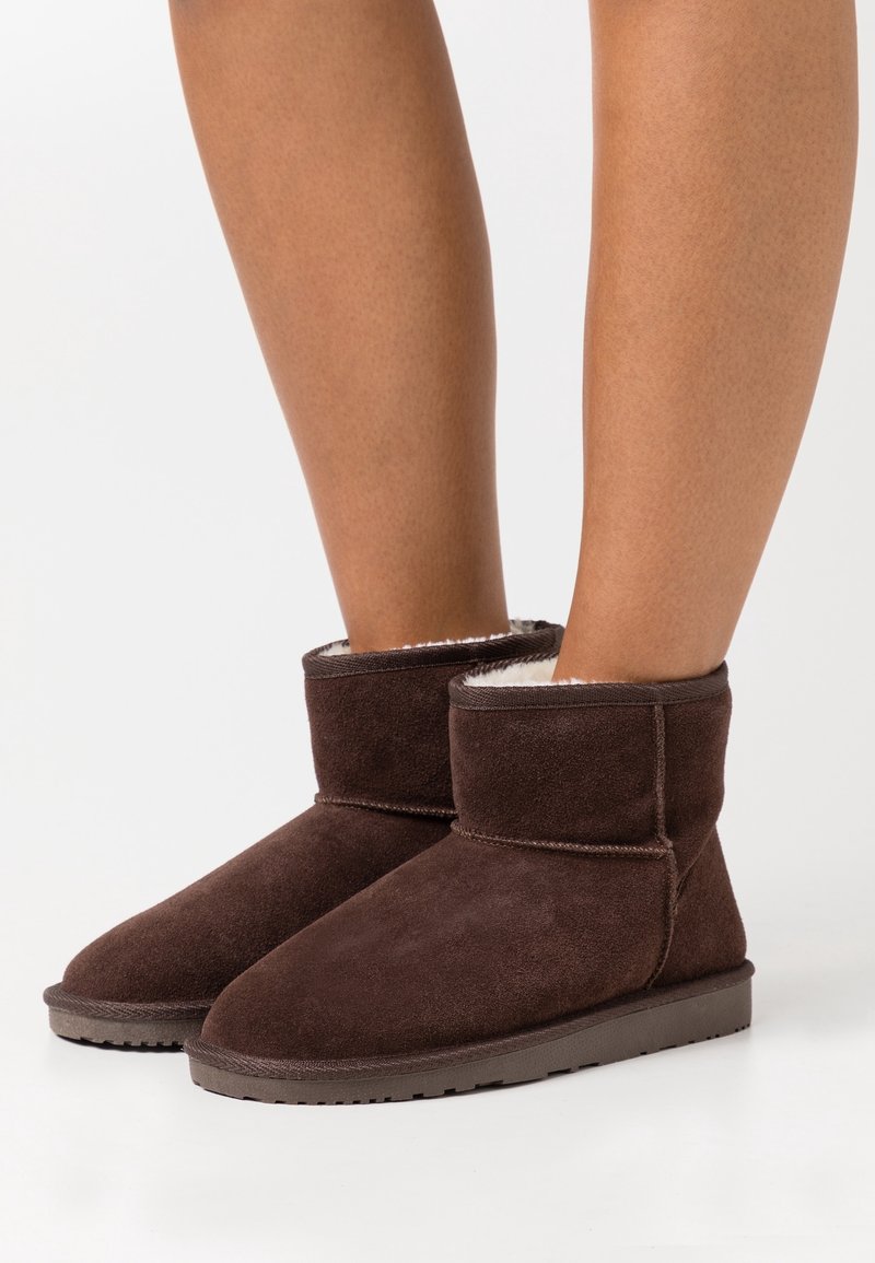 Jambes portant des bottines courtes en daim brun foncé avec une doublure intérieure douce et claire et des semelles en caoutchouc plates sur fond blanc.
