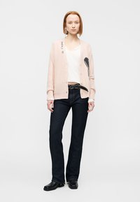 Γυναίκα με ανοιχτό ροζ ζακέτα με μαύρο σχέδιο, λευκό πουκάμισο, μαύρη ζώνη, σκούρο μπλε τζιν και μαύρα παπούτσια, στέκεται μπροστά σε λευκό φόντο.