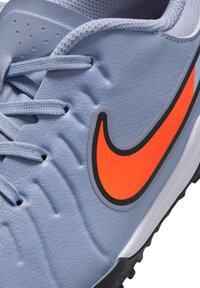 Chaussure de football Nike avec une tige synthétique bleu clair, surface texturée, lacets gris et un logo swoosh orange vif sur le côté. Semelle en caoutchouc pour une meilleure traction.