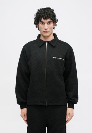 Veste zippée noire avec un motif texturé, dotée d'un col, de manches longues et d'une poche avant avec une fermeture éclair métallique.