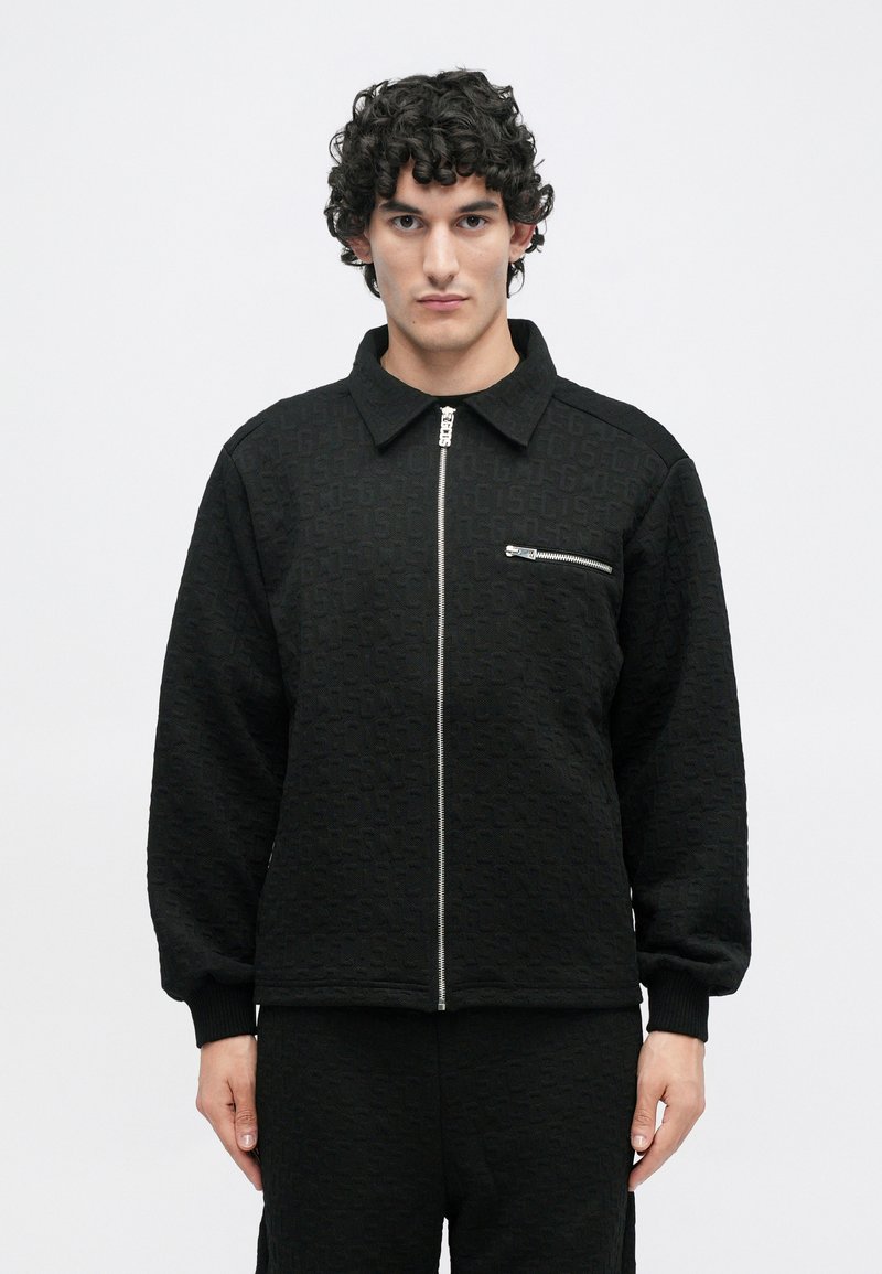 Chaqueta negra con cremallera, con un patrón texturizado, que presenta un collar, mangas largas y un bolsillo frontal con una cremallera metálica.