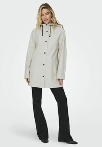 Imperméable beige clair avec une capuche, des boutons noirs et un cordon de serrage. Il présente deux poches latérales et une coupe droite. Porté sur un pantalon noir évasé.