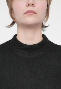 Melnas ribotas adītas džemperis ar augstu kaklu, kura audums ir mīksts un cieši pieguļošs. Audums gludi līdzinās ap kaklu.