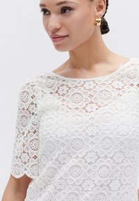 Top bianco in pizzo con motivi floreali, maniche corte e scollatura ondulata. Modella accessorata con orecchini a cerchio dorati.