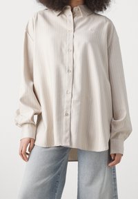 Camicia button-up beige chiaro con sottili righe verticali, maniche lunghe e vestibilità rilassata. Realizzata in tessuto morbido con colletto.