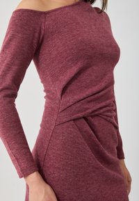 Burgundi színű, off-shoulder ruha texturált kötött anyagból, hosszú ujjakkal és derékon ráncolt részletekkel a fokozott dimenzió érdekében.