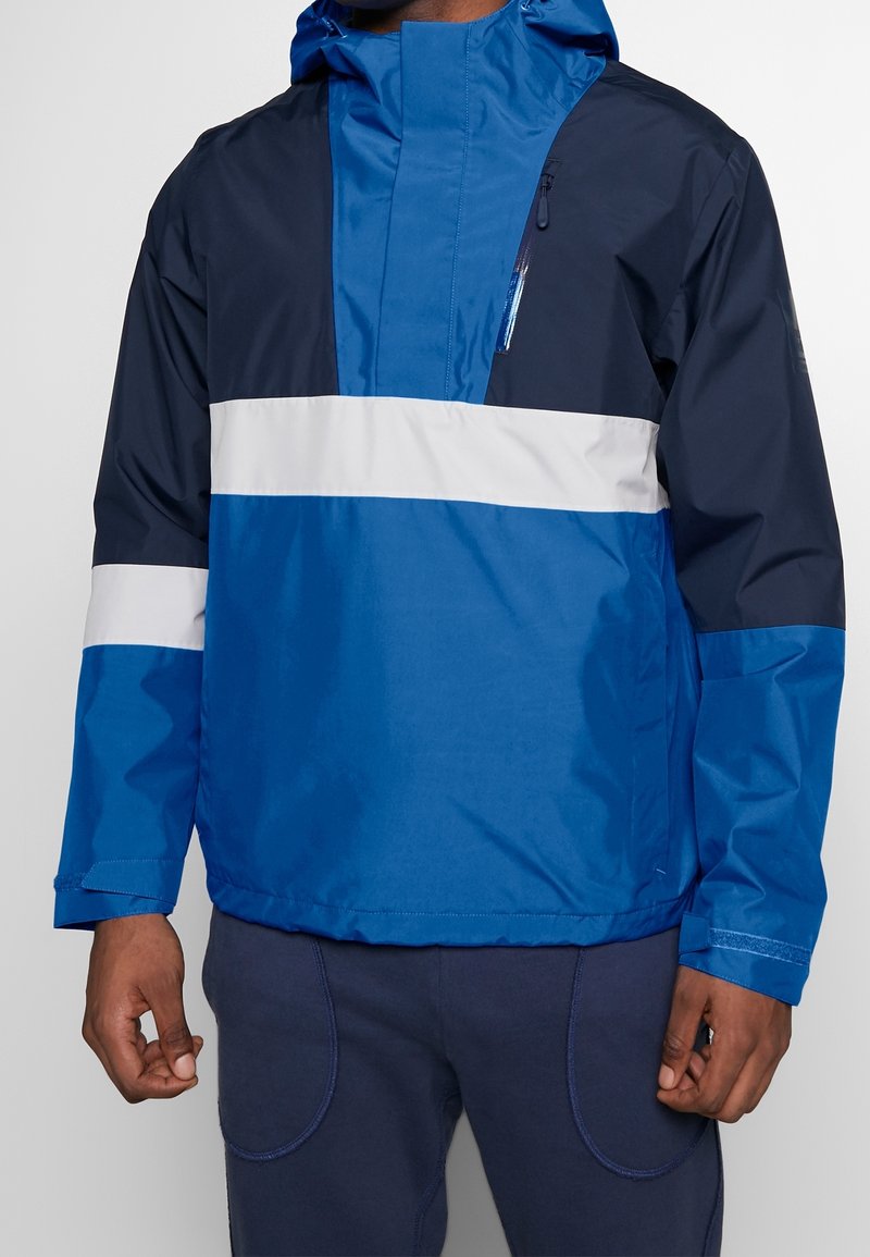 Giacca a vento blu e navy con una striscia orizzontale bianca, tasca con zip frontale e cappuccio regolabile. Materiale liscio e resistente all'acqua.