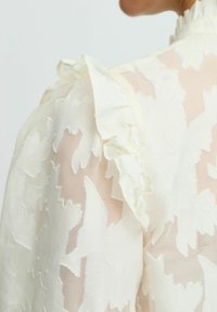 Blusa bianca con motivo floreale e dettagli arricciati sulle spalle. Realizzata in tessuto trasparente, presenta ricami floreali a rilievo.