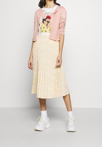 Ljusrosa cardigan över en vit t-shirt med grafik som har en färgglad design, i kombination med en plisserad gul blommig midiklänning och vita sneakers.