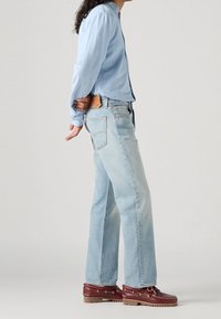 Camisa azul claro abotonada, vaqueros de lavado claro y pierna recta, y zapatos náuticos rojo oscuro con detalles en color canela y suelas marrones.