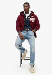 Felpa bordeaux con logo frontale, camicia blu navy sotto, jeans azzurri chiari e sneakers beige con suola bianca, seduto su uno sgabello nero.