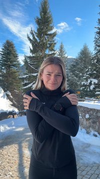 Haut de sport noir à manches longues et coupe ajustée. La personne se tient debout en plein air dans un environnement enneigé, se serrant elle-même dans ses bras.