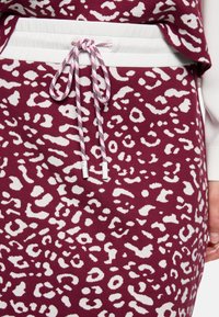 Bordeauxroter Stoff mit weißem Tierdruckmotiv. Der Bund hat eine weiße Kordel mit Knoten und gestreiften Akzenten.