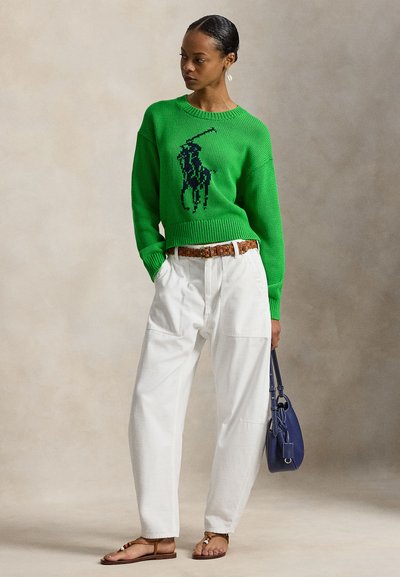 Πράσινο πλεκτό πουλόβερ με μαύρο λογότυπο αλόγου, λευκό φαρδύ παντελόνι, καφέ ζώνη και σανδάλια, παρουσιασμένα πάνω σε απαλό μπεζ φόντο.