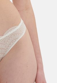 Witte kanten string met intrikate bloemenpatronen, zachte textuur en golvende randen, ontworpen voor een minimale bedekking.