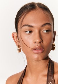 Hnedý horúcik s flitrami; modelka nosí veľké, lesklé zlaté náušnice. Pokožka je hladká s prirodzeným leskom. Vlasy sú stiahnuté dozadu.