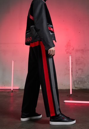 Personne portant une veste en cuir noire et un pantalon large noir avec des rayures verticales rouges et grises, debout sur un sol en béton avec un éclairage rouge.