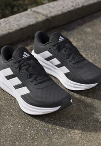 Chaussures de course noires et blanches avec trois bandes blanches sur le côté, posées sur un trottoir extérieur près d’un bord en pierre.
