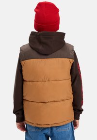 En person i röd mössa, brun hoodie och tan-färgad pufferväst, stående med ryggen mot kameran mot en enkel bakgrund.