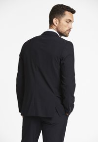 Completo blu marino con taglio su misura, spalle strutturate e spacco singolo nella parte posteriore. Presenta una camicia bianca con colletto sotto.