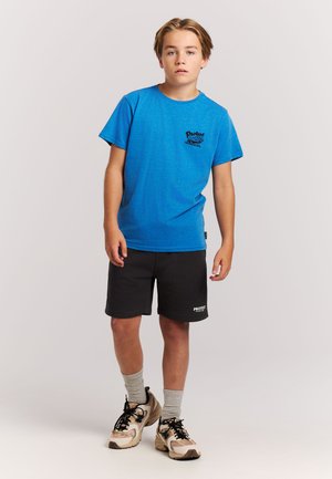 Jonge persoon die staat en een blauw t-shirt, zwarte korte broek, grijze sokken en beige sneakers draagt tegen een effen lichte achtergrond.