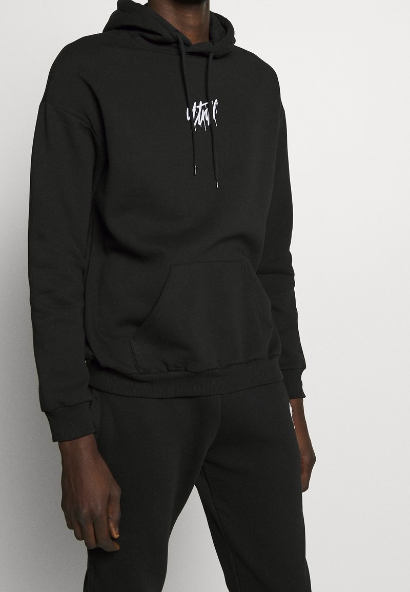 Homme portant un sweat à capuche noir avec un texte blanc en écriture manuscrite et un pantalon de survêtement noir assorti, sur un fond clair uni.