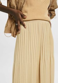 Personne à la peau foncée portant une jupe plissée beige et un haut en maille assorti avec une veste marron clair, main posée sur la taille de la jupe.