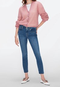 Cárdigan de punto rosa claro con escote en V profundo y botones, combinado con jeans ajustados azul y mocasines blancos con acentos plateados.