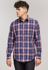 Mężczyzna z kręconymi włosami, ubrany w niebieską i czerwoną koszulę w kratę z guzikami, patrzy w lewo na tle jasnym jednolitym.