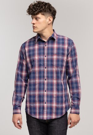 Mężczyzna z kręconymi włosami, ubrany w niebieską i czerwoną koszulę w kratę z guzikami, patrzy w lewo na tle jasnym jednolitym.