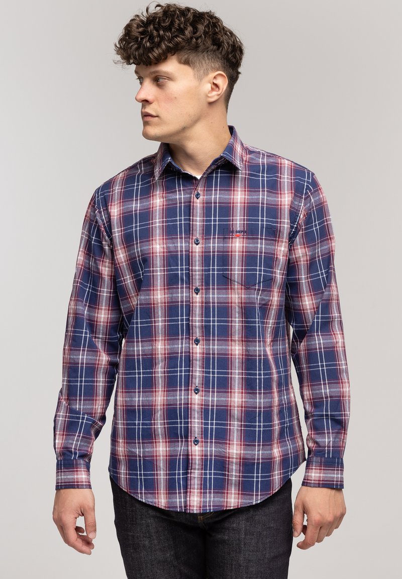 Mężczyzna z kręconymi włosami, ubrany w niebieską i czerwoną koszulę w kratę z guzikami, patrzy w lewo na tle jasnym jednolitym.