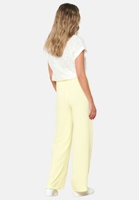 Biała haftowana bluzka z krótkim rękawem, zestawiona z luźnymi, teksturowanymi żółtymi spodniami. Model ma na sobie białe buty. Gładkie tło.