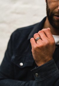 Anello d'argento con motivi intricati, indossato sulla mano destra. La mano è parzialmente visibile, appoggiata contro una giacca scura. Lo sfondo è tenue.