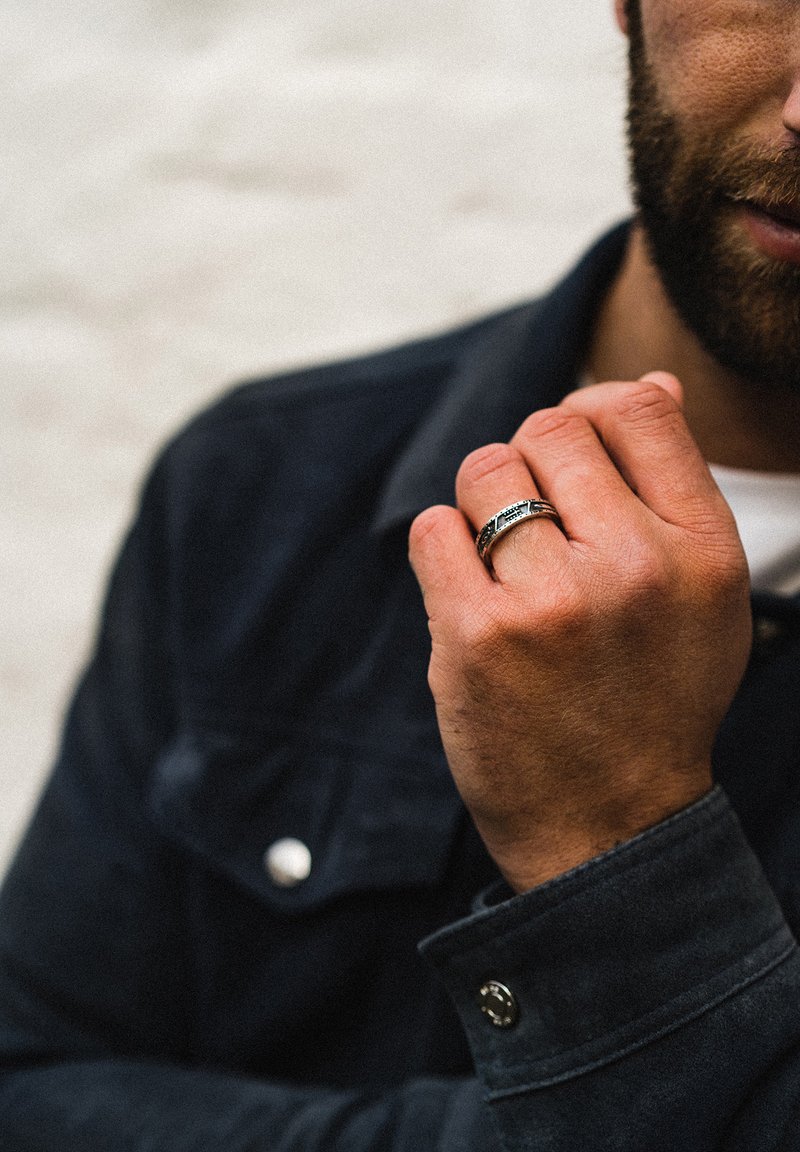 Anello d'argento con motivi intricati, indossato sulla mano destra. La mano è parzialmente visibile, appoggiata contro una giacca scura. Lo sfondo è tenue.