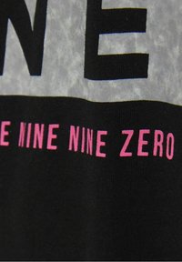 Černá tkanina s velkými bílými písmeny tvořícími „JEDEN“ a menším růžovým textem níže, který uvádí „DEVĚT DEVĚT NULA“, na texturovaném pozadí.