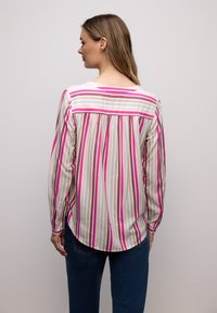 Femme de dos portant un chemisier à manches longues à rayures verticales rose, beige et blanche, associé à un jean bleu foncé sur un fond uni.