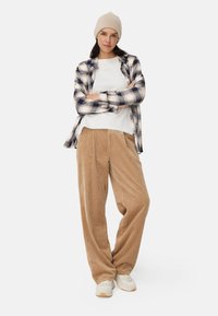 Bonnet crème, chemise à carreaux bleu marine et beige, t-shirt blanc et pantalon en velours côtelé beige avec plis ; des baskets beiges complètent la tenue.