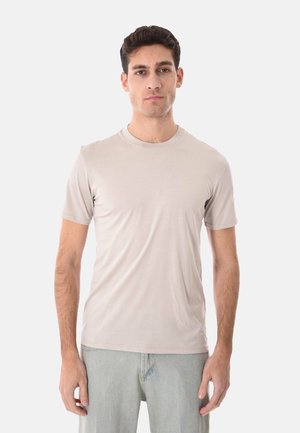 Uomo in piedi di fronte, indossa una maglietta a maniche corte color beige chiaro con scollo rotondo e jeans chiari, su sfondo bianco semplice.