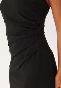 Černé šaty s přiléhavým střihem, které mají zkroucené detaily z látky a vertikální švy, s hladkým, matným povrchem.