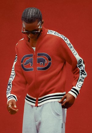 Red zip-up jacket with black and white striped cuffs, featuring a bold graphic design on the front. Model wearing gray sweatpants and sunglasses.