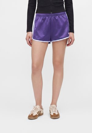 Person steht und trägt violette Sportshorts mit weißem Rand, ein schwarzes Langarmshirt und beige Sneakers mit braunen Streifen.