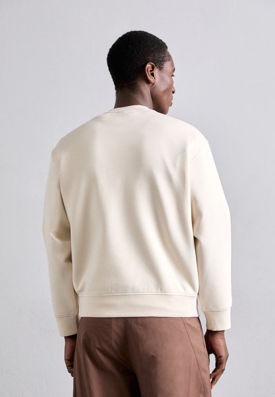 Crèmekleurig sweatshirt met een ronde halslijn, geribbelde mouwen en een zoom. Heeft een zachte textuur en een ontspannen pasvorm. Bruine broek zichtbaar eronder.