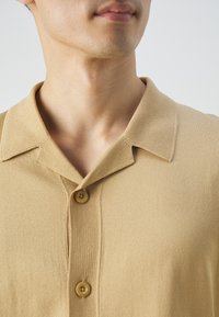 Chemise beige à manches courtes avec un col pointu et deux boutons. Le tissu est texturé avec une finition douce et côtelée.