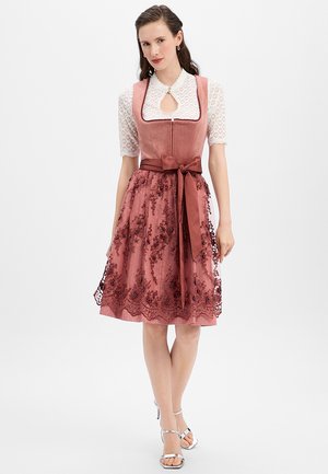 Frau trägt ein knielanges, staubrosafarbenes Kleid mit Spitzenbluse, floraler Stickerei als Überwurf, breitem Taillengürtel und silbernen High-Heel-Sandalen.