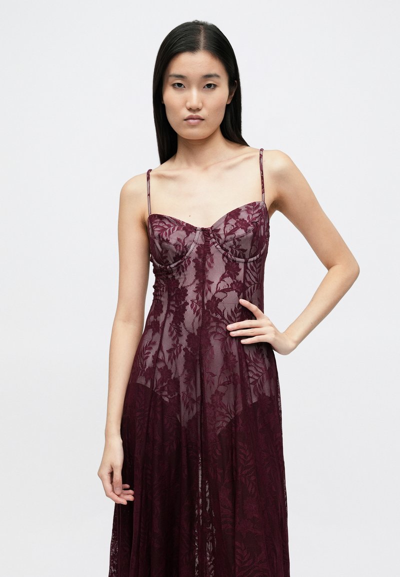 Robe en dentelle bordeaux avec un corsage ajusté, des bretelles fines et une jupe fluide ornée de motifs floraux et d'une texture semi-transparente.
