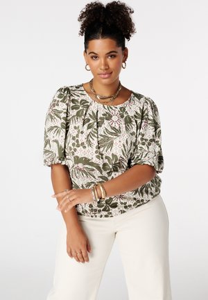 Vrouw met krullend haar in een hoge knot, die een groen-witte bloemenblouse met pofmouwen en een witte broek draagt, geaccentueerd met gouden sieraden.