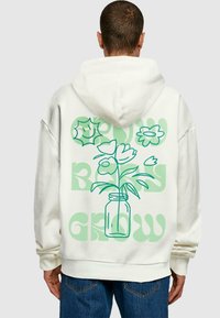 Hvid hættejakke med en blomstergrafik i grøn, med teksten "GROW" og "BLOOM" i fede bogstaver. Blød tekstur og afslappet pasform.