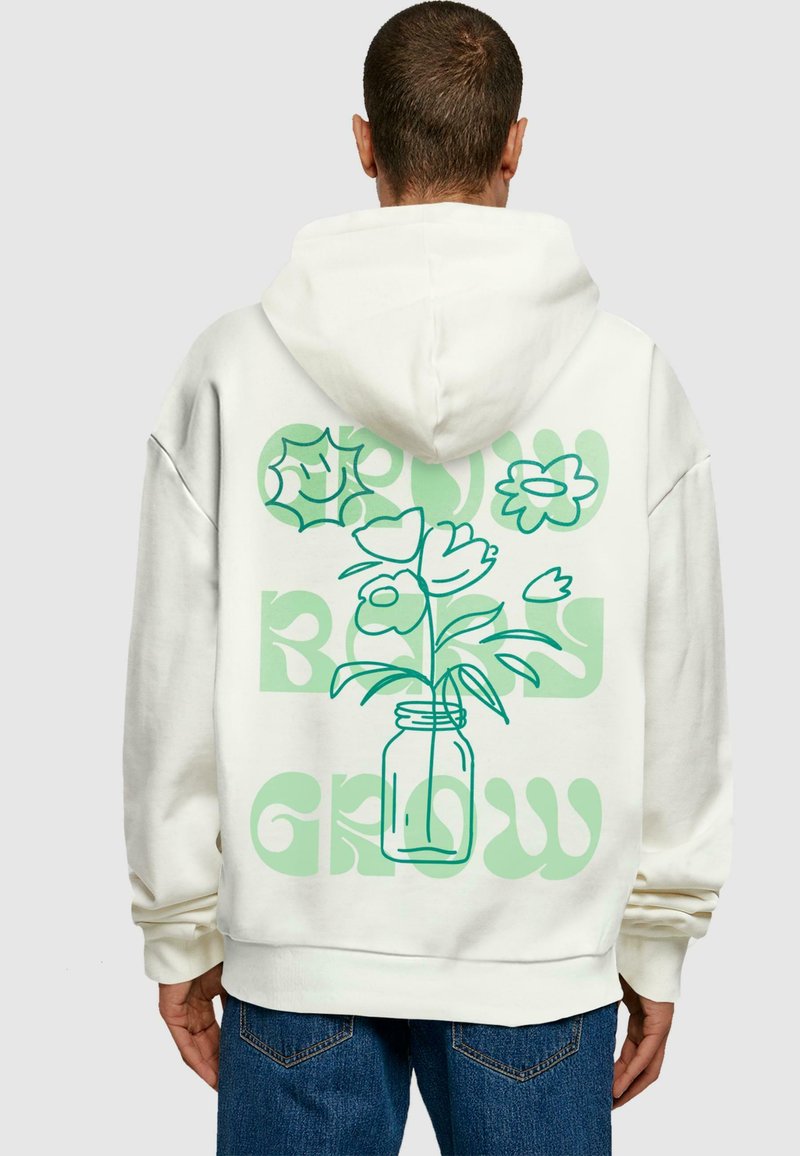 Balta kapuce ar ziedu grafiku zaļā krāsā, ar tekstu "GROW" un "BLOOM" treknrakstā. Mīksta tekstūra un brīva piegriezuma.