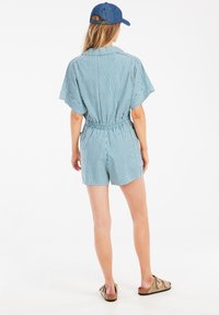 Randig denimromper med korta ärmar, åtdragen midja och shorts. Blå och vita ränder; matchad med en blå keps och beige sandaler.