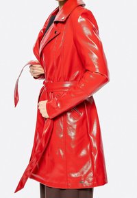 Femme portant une veste en cuir rouge brillante avec ceinture et col à revers, tenant la ceinture près de la taille, sur un fond neutre.